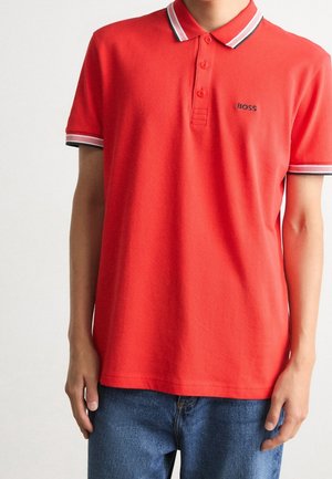 Czerwona koszulka polo z krótkim rękawem, z białym i czarnym obszyciem na kołnierzu i rękawach, noszona z niebieskimi dżinsami, z małym logo „BOSS” na piersi.