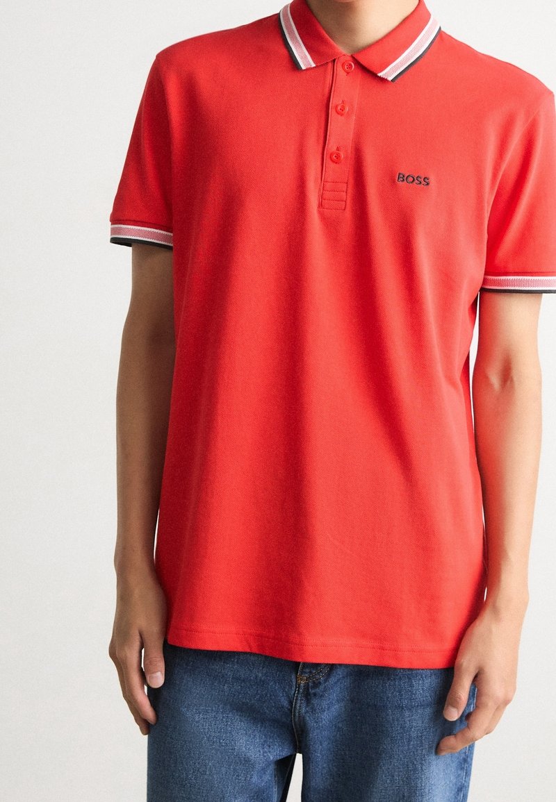 Polo rouge à manches courtes avec des bordures blanches et noires sur le col et les manches, porté avec un jean bleu, arborant un petit logo "BOSS" sur la poitrine.