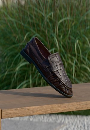Mocassin en cuir marron avec motif crocodile flottant au-dessus d'une surface en bois, herbe verte floue en arrière-plan.