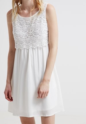 Biała sukienka bez rękawów z misterną koronkową nakładką na górze, luźną spódnicą, noszona przez osobę o falowanych blond włosach na jednolitym tle.