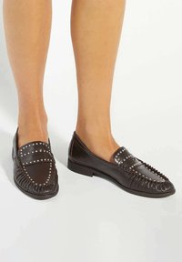 Brown leather loafers with silver studs, detailed stitching, and a flat sole. Features a rounded toe and a gathered fringe along the front.