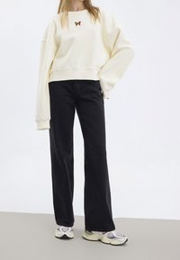 Felpa bianca oversize con ricamo di una farfalla sul petto, abbinata a jeans neri a gamba larga e sneakers bianche sportive.