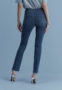 Kitsad teksad tumesinises denimis, keskmise kõrgusega vöökoht, kaks tagataskut ja sobiv siluett, koos valgete kõrge kontsaga sandaalidega.