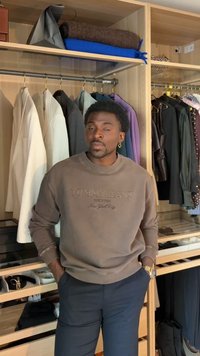 Homme en sweat-shirt marron et pantalon foncé, debout les mains dans les poches devant une armoire en bois avec des vestes suspendues et des vêtements pliés.