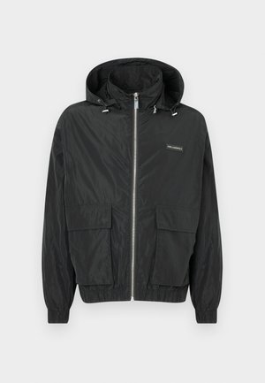 Svart lett jakke med hette, to frontlommer, elastiske mansjetter og en hel glidelås. Har et logoemblem på venstre side.