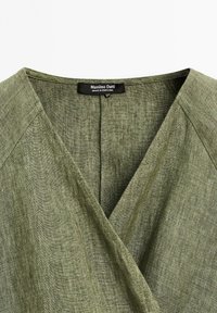 Detailaufnahme des Halsausschnitts eines grünen Leinenkleidungsstücks mit V-Ausschnitt und Markenlabel "Massimo Dutti, Made in Portugal."