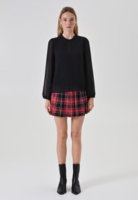 Schwarze Bluse mit langen Ärmeln aus transparentem Stoff und einem Schlüsselloch-Ausschnitt, kombiniert mit einem rot-schwarz karierten Faltenrock. Schwarze Knöchelstiefel runden das Outfit ab.