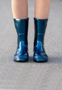 Bottes de pluie en caoutchouc bleu avec finition brillante, modèle jusqu'au genou, semelles texturées et accents de boucle sur le côté, posées sur une surface en béton.