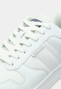 White sneaker featuring a smooth synthetic upper, lace closure, subtle logo tag, and a rounded toe design with tonal stitching details.