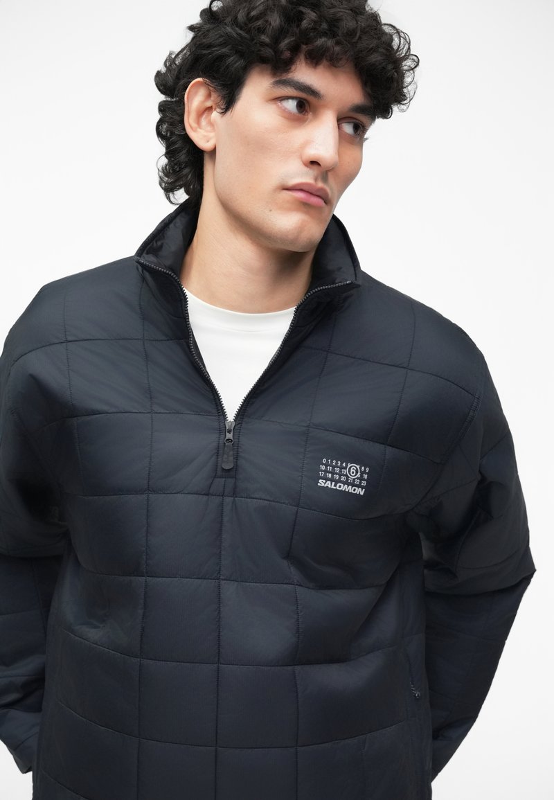 Black quilted pullover jacket with a zippered collar, featuring a front logo and a smooth texture. Lightweight and designed for warmth.