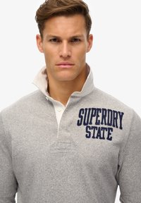 Polo de manga larga gris con un cuello blanco, que presenta el texto "SUPERDRY STATE" bordado en azul marino en el área del pecho izquierdo.