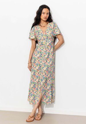 Femme aux longs cheveux foncés portant une robe à fleurs multicolore à boutons sur le devant, manches courtes et sandales marron, debout contre un mur blanc.