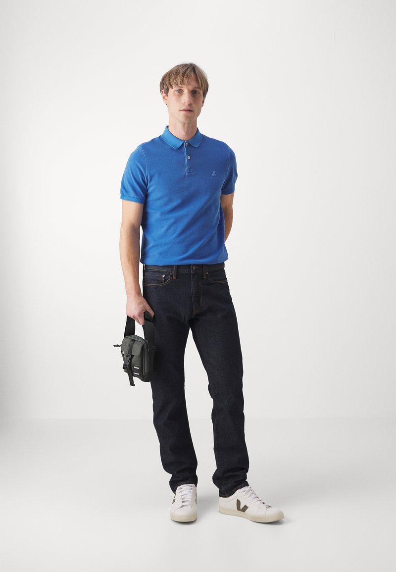 Camisa polo azul, jeans oscuros y zapatillas blancas. Detalle adicional: modelo sosteniendo una pequeña bolsa verde. Diseño simple y moderno.