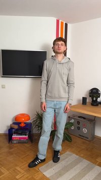 Gray hoodie with kangaroo pocket, light blue jeans, and black shoes with white accents. Standing on a patterned rug in a modern interior.