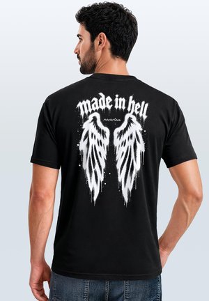 Mann mit dunklen Haaren und Bart, der ein schwarzes T-Shirt mit weißen Engelsflügeln und dem Text "made in hell nevertheless" auf dem Rücken trägt.