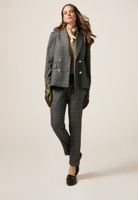 Blazer à carreaux gris avec des boutons croisés associé à un pantalon à carreaux assorti. Le mannequin porte des gants en cuir verts et des chaussures noires.