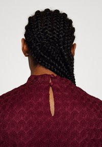 Femme aux cheveux tressés foncés de dos, portant un haut bordeaux texturé avec deux boutons recouverts de tissu au niveau de l'encolure arrière.