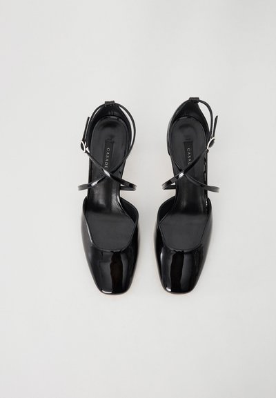 Sorte patentlæder slingback-sko med firkantede tåer, tynde ankelstropper og en mat sort indersål med et logo. Glat overflade.