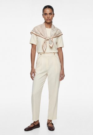 Frau trägt cremefarbenes Kurzarmoberteil und hochtaillierte Hose, gestreifter beiger Schal an den Schultern gebunden und dunkelbraune flache Schuhe, steht aufrecht.