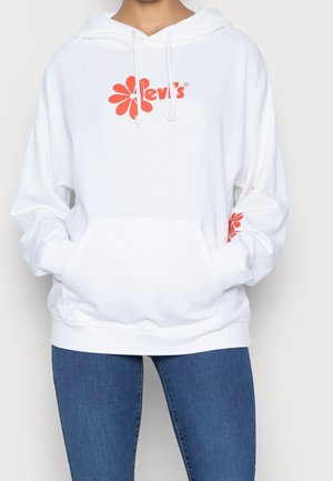 Osoba ubrana w białą bluzę z kapturem Levi’s z czerwonym logo kwiatu na piersi i rękawie, w połączeniu z niebieskimi dżinsami. Ręce wsadzone w kieszeń bluzy z kapturem.