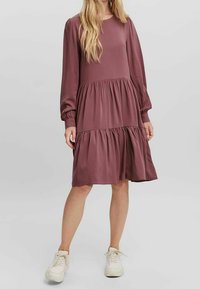 Abito midi a strati di colore viola, con maniche lunghe, tessuto morbido, scollo rotondo e vita arricciata. Modella abbinato a sneakers bianche.