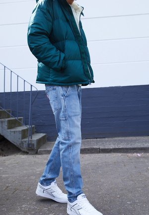 Veste verte rembourrée avec une doublure en polaire blanche, jeans bleu clair avec poches, et baskets blanches, sur une personne debout près des escaliers.