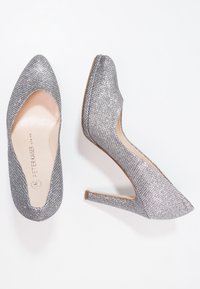 Pár stříbrných třpytivých lodiček na vysokém podpatku s špičatými špičkami, jedna bota zobrazená shora, druhá z boku.