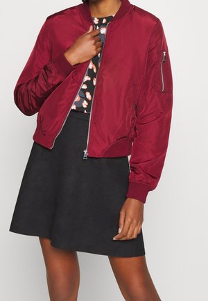 Personne portant une veste bomber rouge sur une jupe noire et un haut noir à motifs, tenant la fermeture éclair de la veste d'une main.