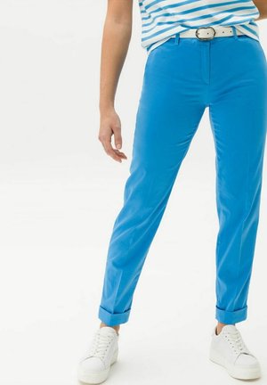 Persoon die felblauwe opgerolde broek draagt, een witte riem, een blauw-wit gestreept topje en witte sneakers, staand tegen een effen achtergrond.