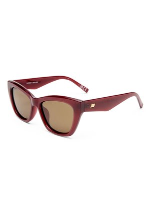 Gafas de sol rojas estilo cat-eye con lentes marrones y montura gruesa y brillante, colocadas en ángulo sobre un fondo blanco.