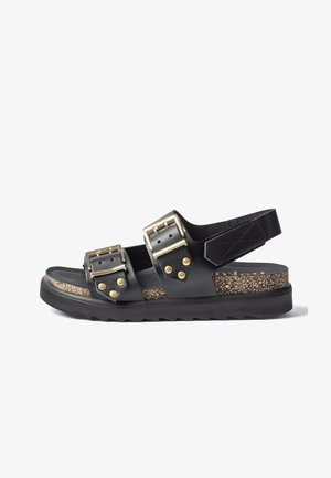 Sandalia de cuero negro con dos hebillas doradas ajustables, plantilla con patrón de corcho, suela negra y una correa de velcro en el tobillo.