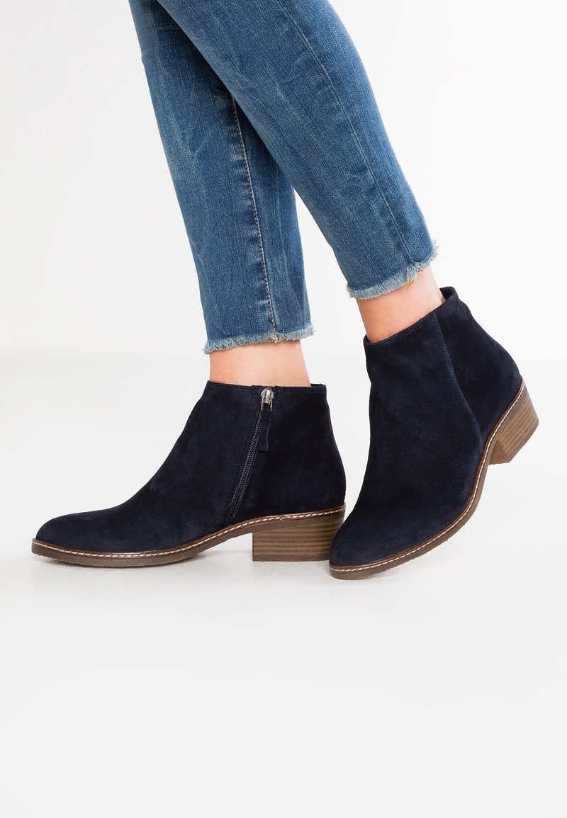 Botas de ante azul marino hasta el tobillo con punta afilada y cremallera lateral. El tacón apilado es marrón, y el diseño es elegante con un detalle mínimo.