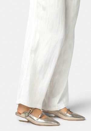 Pies con zapatos de tacón bajo en plateado metálico con puntera puntiaguda y abertura trasera, combinados con pantalones blancos anchos y sueltos con textura.