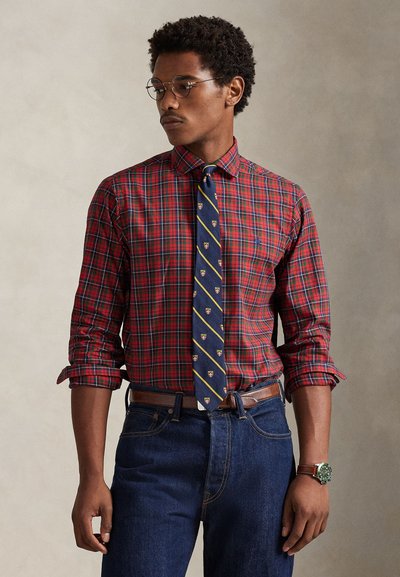 Camicia a quadri rossa e verde con bottoni e maniche arrotolate, abbinata a una cravatta blu scuro con strisce dorate ed emblemi; jeans di denim.