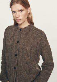 Bruine kabelgebreide cardigan van gestructureerde stof, met een ronde hals, knoopsluiting en opvallende golfpatronen.