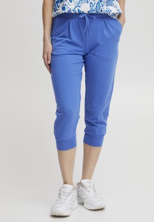 Kvinde iført blå cropped joggerbukser med snøre i taljen og hvide sneakers, stående med hænderne i lommerne.