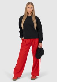 Felpa nera, pantaloni rossi larghi, in mano un cappellino nero. Sneakers con suole marroni. Lunghi capelli biondi lisci. Sfondo grigio neutro.
