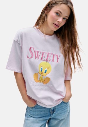 Junge Frau mit langen Haaren trägt ein hellrosa übergroßes T-Shirt mit einem Tweety Bird Motiv und den Texten "SWEETY" und "Leg dich nicht mit diesem Küken an."