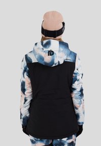 Winterjas in blauwe, zwarte en roze abstracte print. Ontwerp met kap, met een zwart lichaam en mouwen met patroon. Model draagt een roze muts.