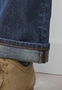 Jeansy denimowe w ciemnoniebieskim odcieniu, z podwiniętymi mankietami ukazującymi jaśniejszy niebieski materiał pod spodem, w połączeniu z beżowymi butami z zamszu.