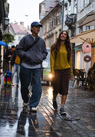 Dva jedinci kráčejí po mokré dlažební ulici. Jeden má na sobě šedou mikinu a modré džíny, zatímco druhý má žlutý svetr a hnědou sukni.