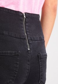 High-waisted black denim jeans with a visible metal zip closure and two back pockets, featuring a smooth texture and understated design.