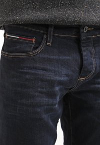 Vaqueros azul oscuro con un corte ajustado, que presentan costuras doradas, un pequeño acento en rojo, blanco y azul en el bolsillo, y herrajes metálicos clásicos.