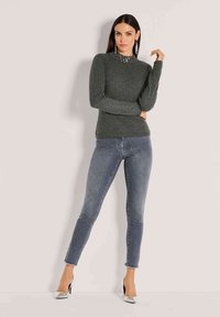 Frau posiert in einem grauen Longsleeve-Oberteil mit gemustertem Kragen, eng anliegenden blauen Jeans, silbernen High Heels und hängenden Ohrringen vor einer schlichten Wand.