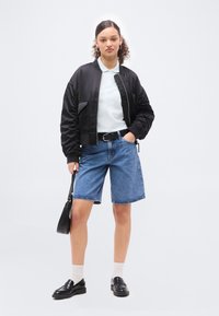 Νέα γυναίκα στέκεται φορώντας μαύρο bomber μπουφάν πάνω από λευκό πόλο πουκάμισο, τζιν σορτς, μαύρη ζώνη, λευκές κάλτσες, μαύρα loafers, κρατώντας μαύρη τσάντα χειρός.