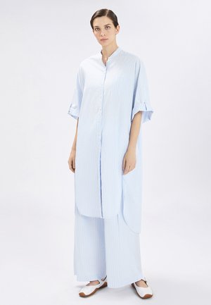 Donna in piedi che indossa una camicia azzurro chiaro lunga con bottoni, maniche arrotolate e pantaloni a gamba larga abbinati, abbinati a scarpe bianche.