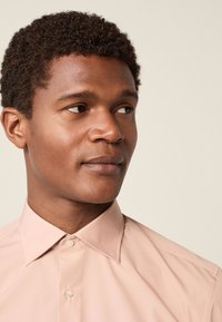 Svijetlo ružičasta košulja sa špicastim ovratnikom i zatvaranjem na gumbe. Glatka tekstura tkanine, uski kroj, naglašen urednim završenim šavom.