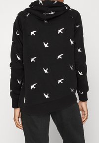 Sudadera negra con una tela texturizada, que presenta patrones de pájaros en plata metálica dispersos por toda la prenda. Incluye una capucha con cordón y puños acanalados.