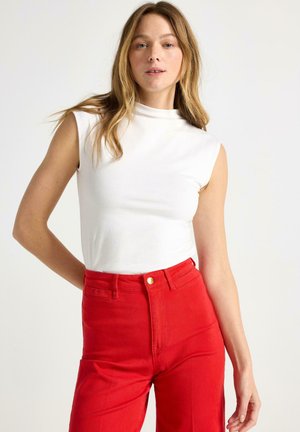 Ung kvinde med langt blondt hår iført en ærmeløs hvid top og rød bukser med høj talje, stående mod en ensfarvet baggrund.