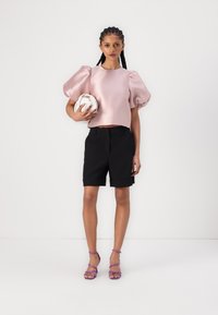 Rosa sateng puff-sleeve topp, svarte skreddersydde shorts, sølv rund clutch og lilla sandaletter med stropper, stående mot en hvit bakgrunn.